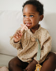 A young child in a patterned beige outfit sits on a couch, smiling slightly while a mushie Flower Training Toothbrush peeks out from under the blanket draped over their legs. The child holds a pink pacifier and looks up thoughtfully.