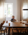 A young child with blond hair and glasses sits at a wooden dining table using mushie Dinnerware Cups among books and art supplies, surrounded by chairs in a sunlit modern room with large windows and light curtains.