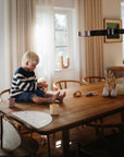A young child with blonde hair and glasses sits on a wooden dining table in a sunlit room, wearing a striped sweater. mushie Dinnerware Cups and decorative items are scattered around them.