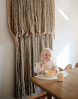 A young child with blonde hair eats at a wooden table from a mushie Round Dinnerware Bowl, BPA-free and yellow, with a matching cup nearby. Sunlight streams past a neutral woven wall tapestry in the background.