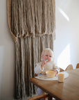 A young child with blonde hair sits at a wooden table, eating from a yellow bowl using the mushie Dinnerware Fork and Spoon Set. A dishwasher-safe beige cup and placemat are on the table as sunlight streams beside a large wall hanging.