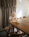 A young child with light blonde hair sits at a wooden table, using mushie Dinnerware Cups and a bowl. Sunlight creates soft shadows across the scene, and a textured wall hanging is visible in the background.