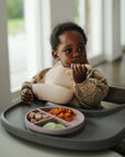 A young child sits in a high chair, wearing a bib and eating pasta from the mushie Silicone Suction Plate. The softly lit background features large windows.