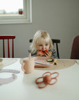 A young blonde child sits at a table eating watermelon. Plates, a sippy cup, a teether, and a mushie Silicone Placemat are on the table. A window and red chair are visible in the background.