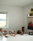 Four young children enjoy eating and drawing around a white table with mushie Dinnerware Cups. The bright room features modern decor, a gold pendant light, and a colorful painting on the wall.