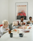 Four young children sit around a white table, enjoying snacks with mushie Silicone Placemats, pastel plates, and cups. A colorful poster decorates the wall behind them as natural light shines in from a window on the left.