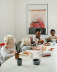 Four young children sit around a white table, enjoying their meal with mushie Square Dinnerware Plates. The colorful plates are neatly arranged, sunlight streams in through the window, and framed artwork decorates the wall.