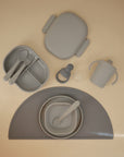 The mushie Lunchbox set features gray non-toxic plastic baby feeding items—divided plate, utensils, sippy cup, lidded container, bowl with spoon, and silicone placemat—arranged neatly on a beige surface.