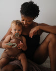Woman sits holding baby with FRIGG Daisy Natural Rubber Pacifier 2-Pack.