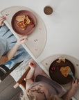 Two young children in light clothing and bibs enjoy heart-shaped pancakes and raspberries at a table, sipping from mushie Dinnerware Cups—creating a charming breakfast scene captured from above.