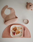 A mushie Dinnerware Cup is shown with a pink silicone bib and matching plate on a placemat. The plate holds heart-shaped pancakes, banana slices, raspberries, and syrup, all neatly arranged on a white table.