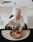 A toddler with curly blonde hair eats from a divided plate at a table featuring a mushie Silicone Placemat made from food-grade silicone, decorated with colorful animal shapes, in a bright kitchen.