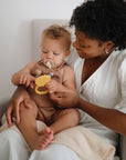 Woman smiles at baby holding Mushie Animal Ring Teether while they sit together on a chair.