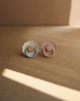 The FRIGG Daisy Silicone Pacifier 2-Pack by FRIGG features one blue and white, and one pink and mauve pacifier, each with a daisy-shaped shield and medical grade silicone nipple, displayed on a brown surface in soft natural light.