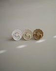 Two daisy-shaped FRIGG Daisy Silicone Pacifiers by FRIGG, in white and beige, are displayed side by side on a neutral surface. Each features a medical grade silicone nipple with soft lighting and gentle highlights in the background.