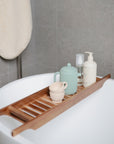 A Mushie Silicone Tea Play Set in mint green sits on a wooden bath tray across a white tub, featuring a beige cup, teapot, and bottles. A beige towel hangs nearby on the gray tiled wall.