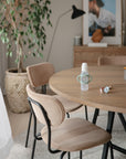 A wooden dining table with beige chairs features a baby bottle fitted with mushie Silicone Bottle Handles and a pacifier on top. In the softly lit background, there’s a potted plant, lamp, and wall art creating a cozy atmosphere.