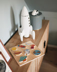 A child’s playroom sideboard features a Mushie Wooden Puzzle, a white toy rocket, a small toy car on a tin, and framed pictures.