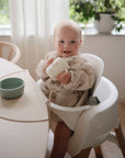 A smiling blonde baby in a beige outfit wears the mushie Long Sleeve Bib while sitting in a high chair, holding a white cup beside a green bowl. Green plants can be seen near the window in the background.