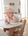 A baby in a high chair, smiling, wearing a cap and holding a mushie HexaPop Suction Toy.