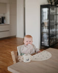 A baby sits in a high chair at a wooden table in a modern kitchen, with one hand near their mouth. In front is the mushie Snack Cup—a spill-proof silicone snack cup perfect for toddlers. Glass cabinets and appliances are visible behind.
