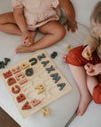 Two young children sit on a white couch playing with a wooden alphabet puzzle. Colorful letters are placed on the board and scattered around them as they interact with the pieces.