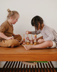 Two young children play with the Mushie Wooden Multi Shape Stacker on a wooden table. One wears a brown outfit, the other a striped shirt and light shorts, focused as they build fine motor skills with this educational toy.
