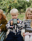 Three kids outdoors: two play with mushie’s Silicone Sensory Toy, one drinks from a mug.