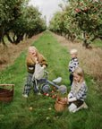 Three kids pick apples with baskets and a mushie Mini Toddler Bike on the grass nearby.