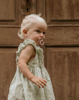 A young blond child in a light dress stands by a rustic brown door, looking to the side with a FRIGG Natural Rubber pacifier from the FRIGG 6-Pack in their mouth.