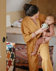 A woman in a mustard yellow robe helps a smiling baby drink from their mushie Bluey Gift Set sippy cup in a cozy, sunlit bedroom with a colorful quilt on the bed.