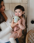 A woman holds a smiling toddler sipping from a green cup with a straw. The toddler, in pink, enjoys her mealtime with the mushie Bluey Gift Set. They are indoors by a wooden chair, and the woman wears a white sweater.