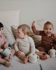 Three toddlers in earth-toned pajamas sit on a bed with neutral bedding. Two hold sippy cups and a mushie Bluey x Mushie Snack Cup—one smiles with a raised hand as they playfully interact with their feeding essentials.