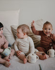 Three toddlers sit on a bed with cream pillows, each using the mushie Bluey Gift Set. One in brown waves and smiles, another in cream looks to the side, and the third in pink gazes at their Bluey silicone baby mealtime cup.