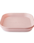 Two light pink, square-shaped mushie Square Dinnerware Plates with rounded edges are stacked together. Made from BPA-free plastic, these stylish and safe plates feature the mushie logo embossed in the center of the top plate.