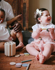 Two toddlers with wooden toys; one uses a mushie Linen Pacifier Clip 2-Pack. Adult in background.