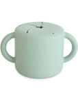 The mushie Snack Cup in Cambridge Blue is a silicone toddler snack cup with two side handles and a flexible, petal-shaped spill-proof lid to prevent messes.