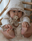 A baby in a textured cream hooded outfit with pom-pom trim sits on a fluffy surface, holding its feet and looking ahead, with a FRIGG Natural Rubber pacifier from the FRIGG 6-Pack clipped to its clothing.