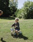A child rides a mushie Mini Toddler Bike on grass as an adult reaches out on a sunny day.