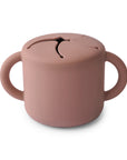 The mushie Snack Cup in Cloudy Mauve is a silicone, spill-proof cup with two handles and a flexible, slit lid that lets little hands reach snacks while minimizing messes.