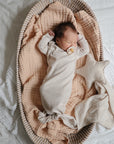 A baby sleeps in a cream knotted gown with a pacifier on a beige quilt in a wicker basket. Nearby, the mushie Star Lovey Blanket is nestled beside the baby, along with a soft star-shaped pillow.