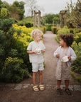 Two toddlers stand on a garden path surrounded by greenery and yellow flowers, smiling at each other while holding mushie Snack Cups. The sky is overcast, and both kids look happy with their spill-proof silicone cups.