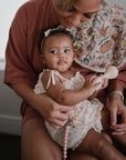 A woman in pink holds a baby girl on her lap. The baby, in a light floral dress and bow headband, grasps a mushie Silicone Baby Rattle Toy made from food-grade silicone. A floral bib rests on the woman's shoulder.