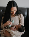A woman with long dark hair sits on a couch, smiling at a baby wrapped in a blanket as she holds the mushie Silicone Baby Rattle Toy, offering the infant a safe and soothing sensory experience.