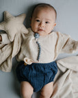 A baby lies on a soft surface, wearing a cream long-sleeve top and dark blue shorts, holding a pacifier with a beaded clip and snuggled up with the mushie Star Lovey Blanket made from organic cotton.