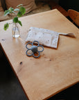 A small wooden table displays a glass jar with a leafy branch, a mushie Water Resistant Wet Bag in white with a subtle pattern for diaper storage, and a gray flower-shaped teething toy in a cozy indoor setting.