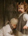 A young girl with curly hair holds a FRIGG Natural Rubber 6-Pack pacifier, standing beside a blond child who looks down. Both wear neutral-toned outfits in front of a wooden background.