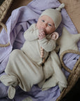 A baby wearing a beige hat and gown lies in a woven basket lined with an organic cotton blanket, holding a beaded pacifier clip, next to the mushie Star Lovey Blanket.