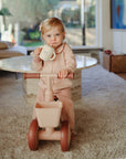 Toddler in pink pajamas on a trike holds a mushie Bluey Gift Set mug on a carpeted floor.