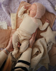 A baby in a pink knit bonnet and beige clothes rests on the mushie Star Lovey Blanket, holding a pacifier as an adult gently supports them. Nearby are a tube of lotion, a star-shaped pillow, and other soft, neutral-toned baby items.